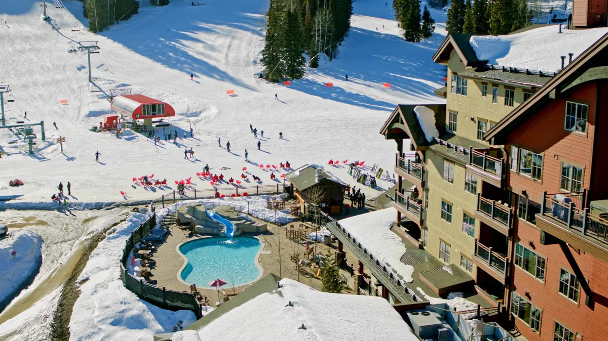 An energetic exterior shot featuring ski slopes, a sparkling outdoor swimming pool, comfortable seating areas, and a ski lift. The snowy environment is alive with activity, showcasing both winter sports and relaxing resort amenities.