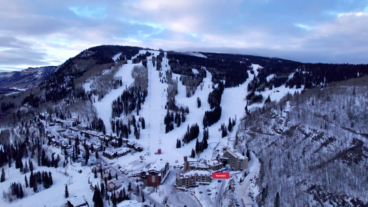 A panoramic view of ski slopes descending through forested hills, framed by a dramatic mountain range. Resort buildings nestle near the slopes, enhancing the sense of a true ski-in, ski-out experience.