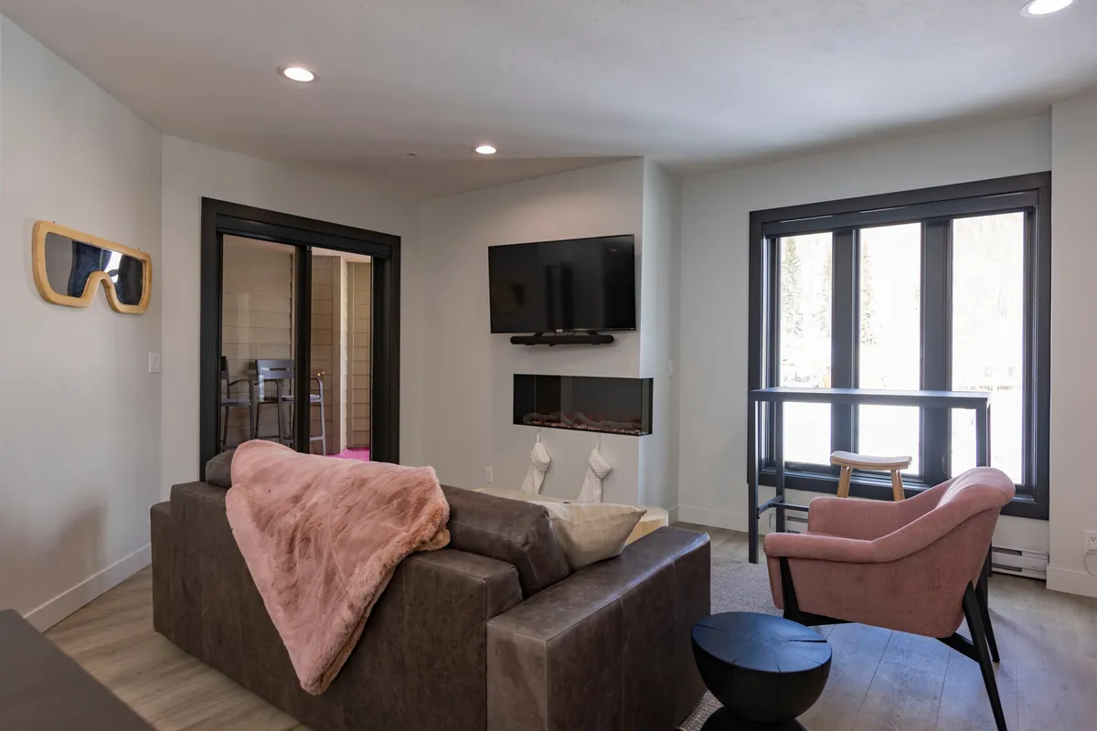 A cozy living area featuring a comfortable sofa with a pink throw blanket, a television, large windows that let in natural light, and playful wall decor. The setting is inviting and relaxed, perfect for unwinding or casual work-from-home moments.