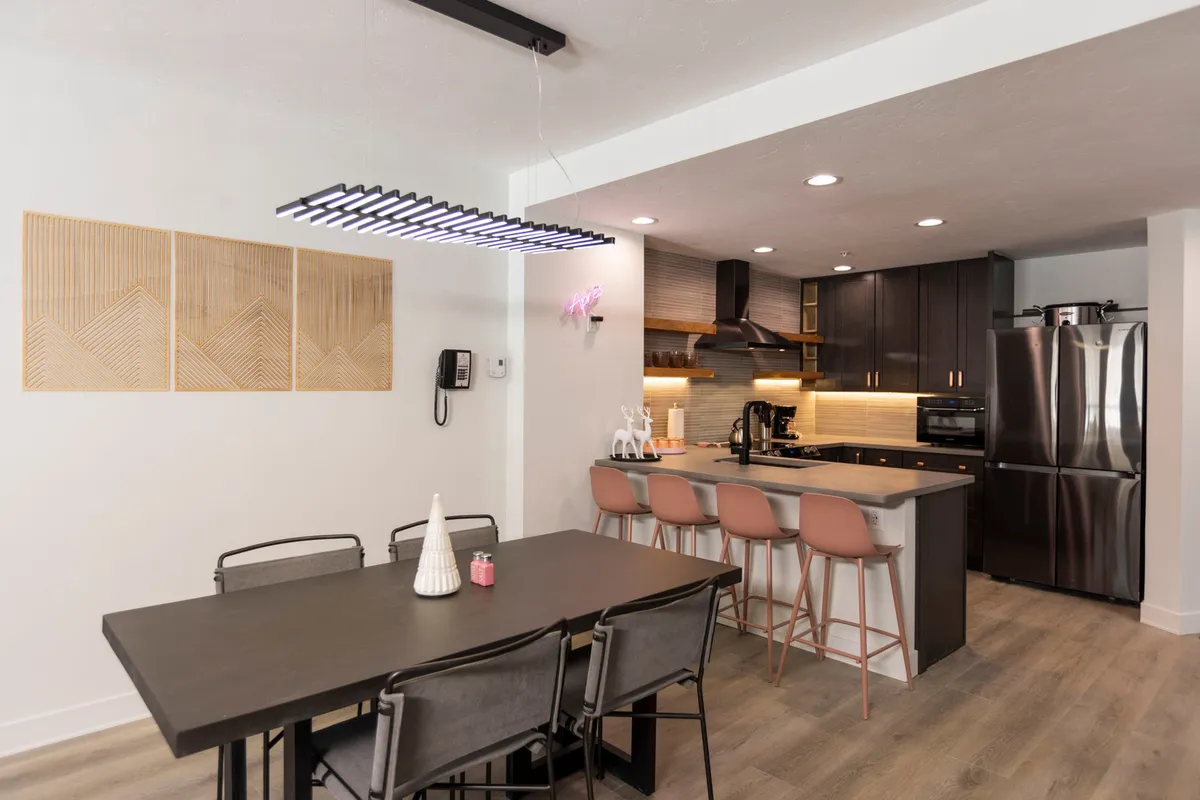 The image showcases a spacious, light-filled dining area with a large wooden table set for gathering, a modern kitchen island equipped for meal prep and socializing, stylish pendant lighting that casts a warm glow, and tasteful mountain-inspired wall art—all coming together to create a contemporary, inviting atmosphere ideal for families and friends.