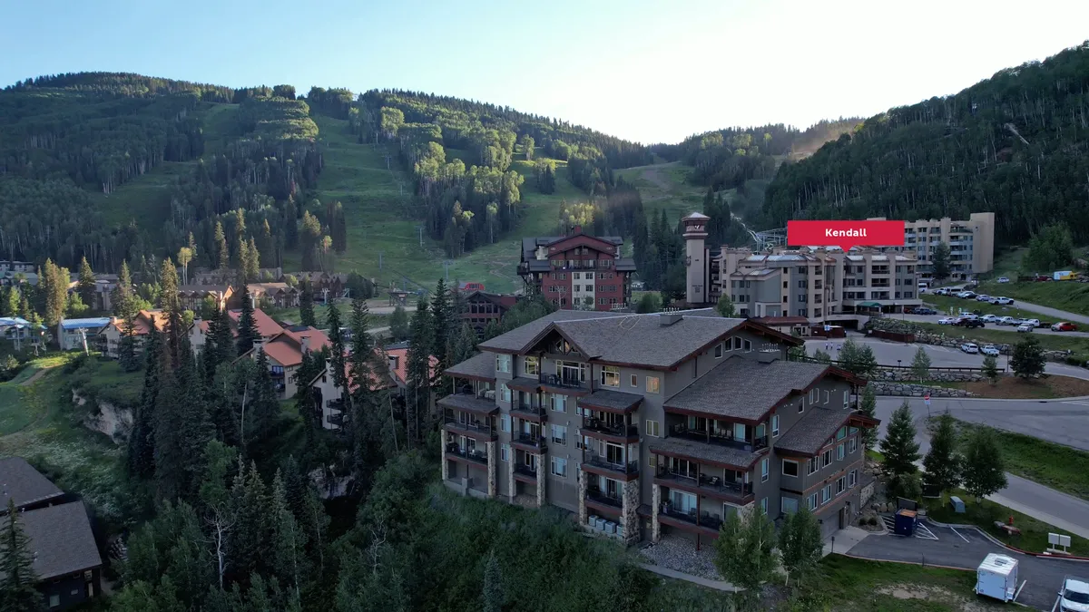 A sweeping exterior view showing mountain lodging buildings nestled at the base of majestic snow-capped mountains. The foreground includes a parking area, while the backdrop features rolling peaks and open skies, capturing the essence of a mountain resort community.