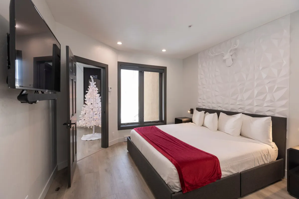 A warmly decorated bedroom featuring a comfortable bed with a plush red throw blanket, a decorative wall with a stag head, and a white Christmas tree in the corner. The room is softly lit, evoking a sense of coziness and festive tranquility.