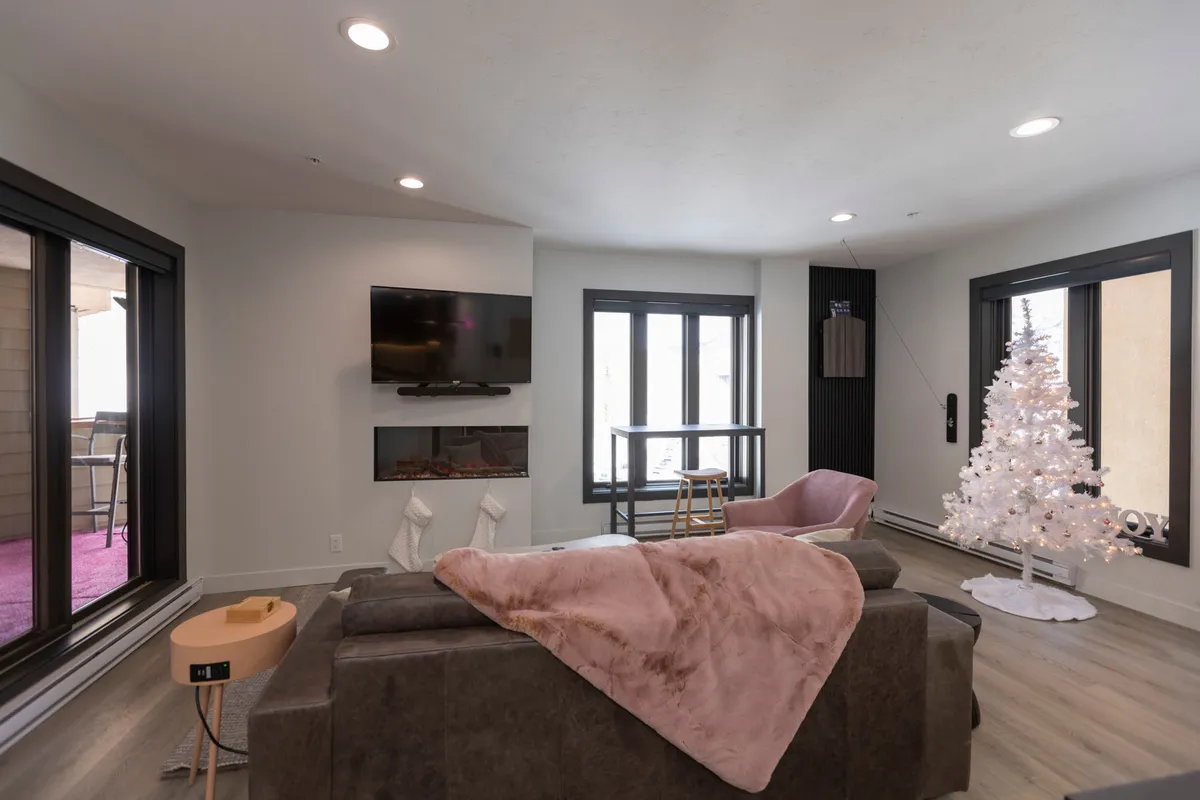 A warm and inviting living room featuring a modern sofa, a crackling wood-burning fireplace, a white Christmas tree, and large windows that fill the space with natural light. The setup is perfect for gathering with family, sharing stories by the fire, and celebrating special occasions together.