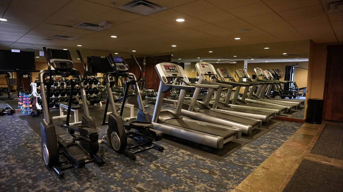 A modern fitness center is shown with elliptical machines, treadmills, dumbbells, mirrors, and exercise mats. The space is well-lit, motivating, and designed for a variety of workout routines, from cardio to strength training and group classes.