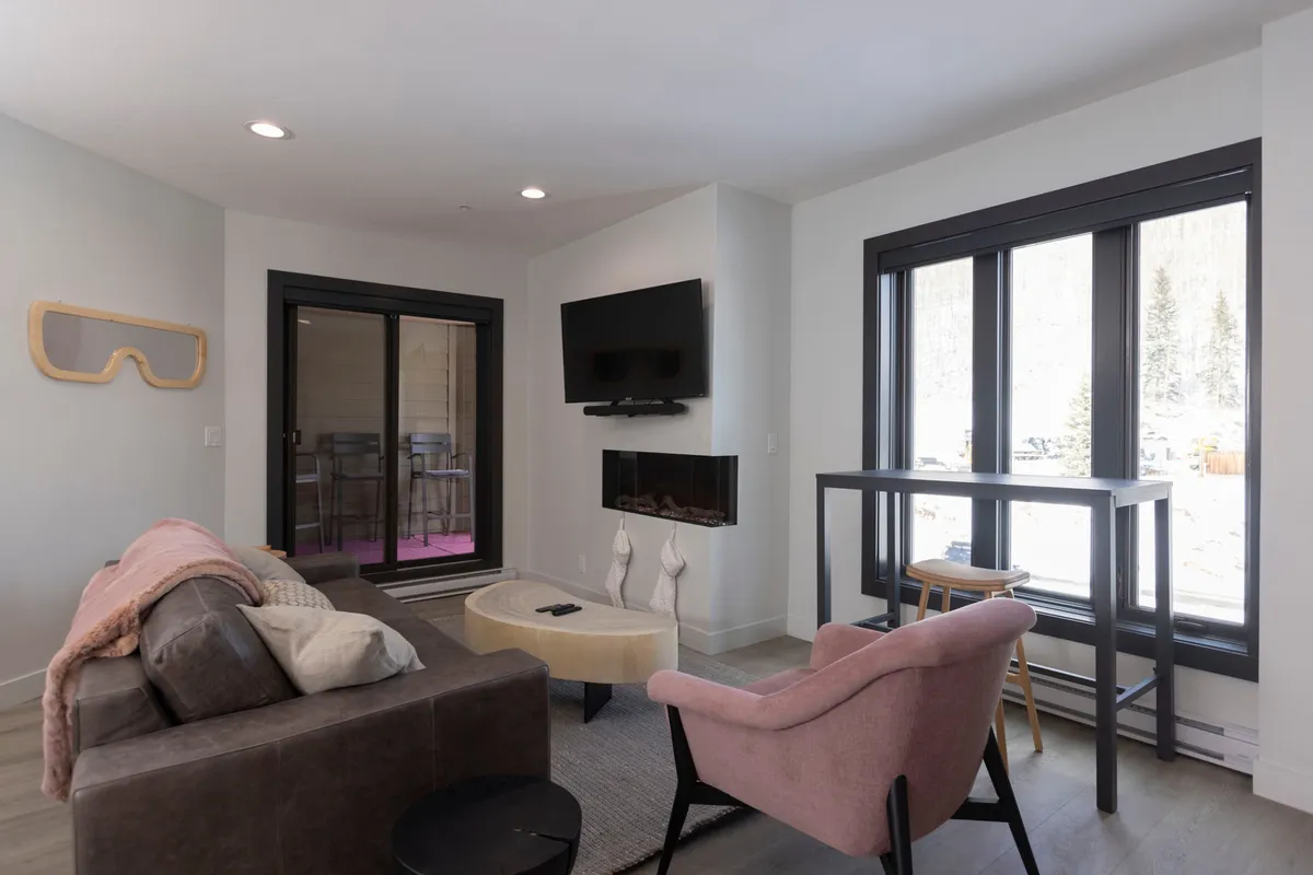 A cozy lounge area featuring a sofa with plush cushions, a pink accent chair, a fireplace, and large windows that bring in abundant natural light and frame glimpses of the outdoors. The inviting setup is perfect for quiet relaxation or enjoying a morning coffee while soaking in the serene views.