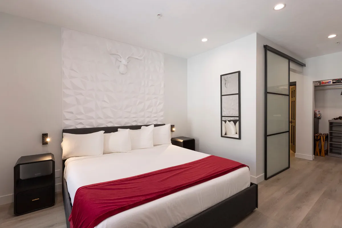 A contemporary bedroom with a bed dressed in a red accent blanket, accompanied by geometric wall art, a stylish decorative wall mount, and black nightstands. The room has a calm, uncluttered atmosphere ideal for relaxation and unwinding.