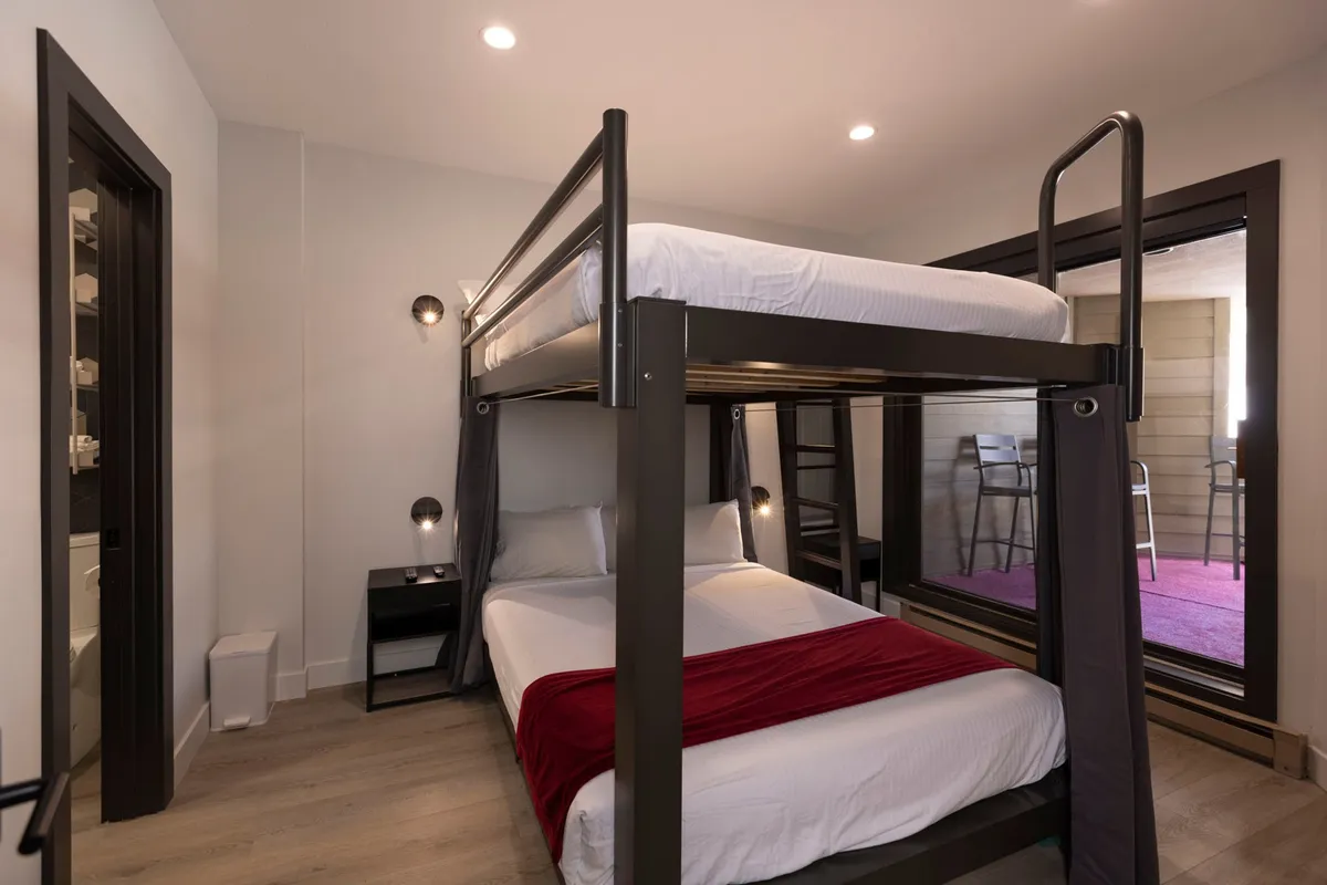 A playful bedroom with a bunk bed dressed in a cozy red blanket, a nightstand, and a window offering natural light. The mood is inviting and warm, supporting restful nights and shared moments for families or friends.