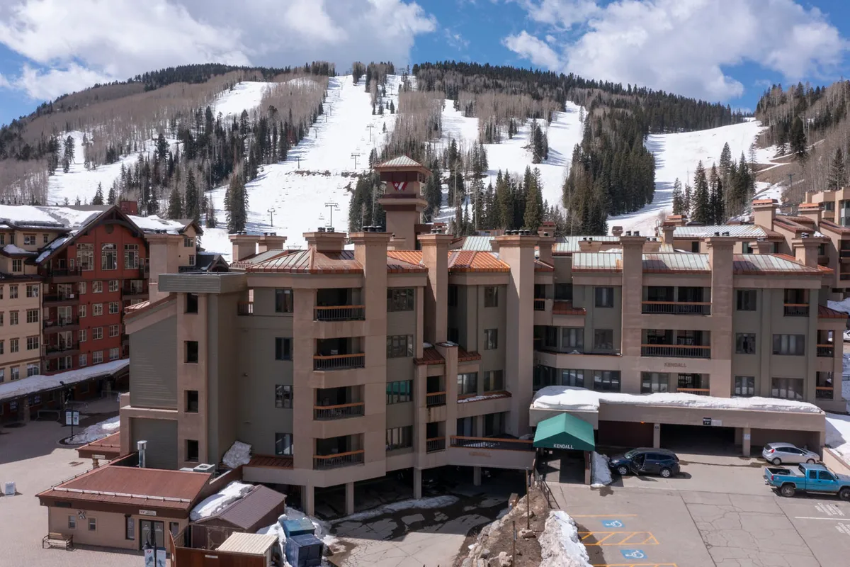 This exterior image showcases the architectural details of the resort building with the mountain slopes in the background and a parking area in the foreground. The scene radiates an adventurous but cozy atmosphere, connecting the property with its prime mountain village location.