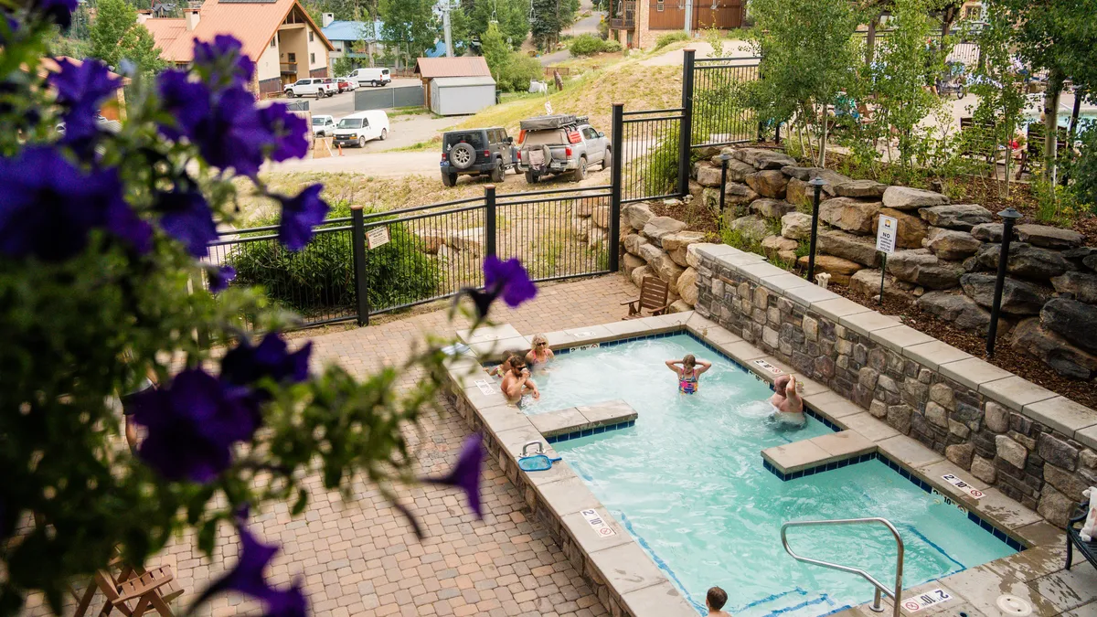 The scene captures a heated outdoor swimming pool surrounded by colorful flowers and dramatic mountain scenery. Guests lounge and socialize poolside, creating a joyful, leisurely atmosphere. The image is set during daylight, emphasizing relaxation and the social aspect of the resort’s amenities.