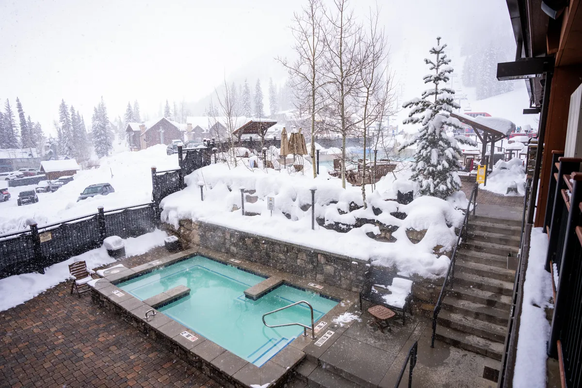 This exterior image features a rooftop hot tub surrounded by a snow-covered landscape and towering trees. Steps lead down from the tub, inviting guests to soak and relax while taking in the serene winter scenery. The mood is tranquil and inviting, perfect for après-ski relaxation.
