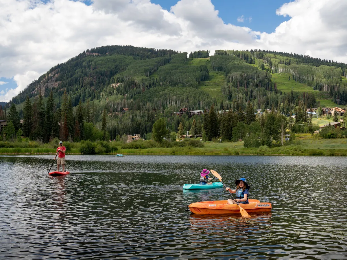 A lively scene featuring kayakers paddling on a reflective body of water with majestic mountains in the background. The image conveys the excitement of family outings and the opportunity for memorable shared experiences in nature, especially during the summer season.