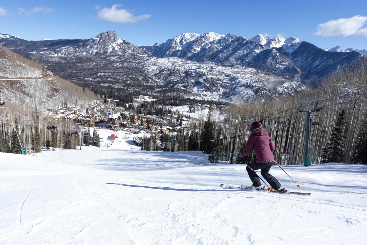 A skier is pictured descending a snowy mountain slope, with a stunning village nestled below and dramatic mountain peaks rising in the background. The sense of excitement and activity is palpable.